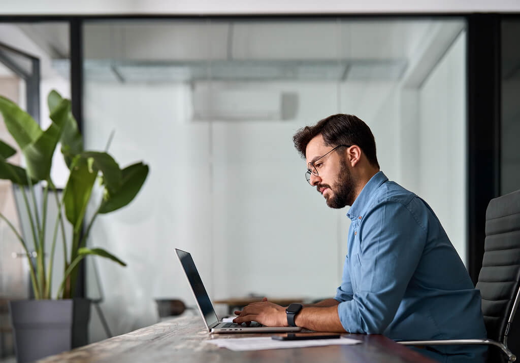Man working on a laptop at a desk in a modern office with plants in the background.