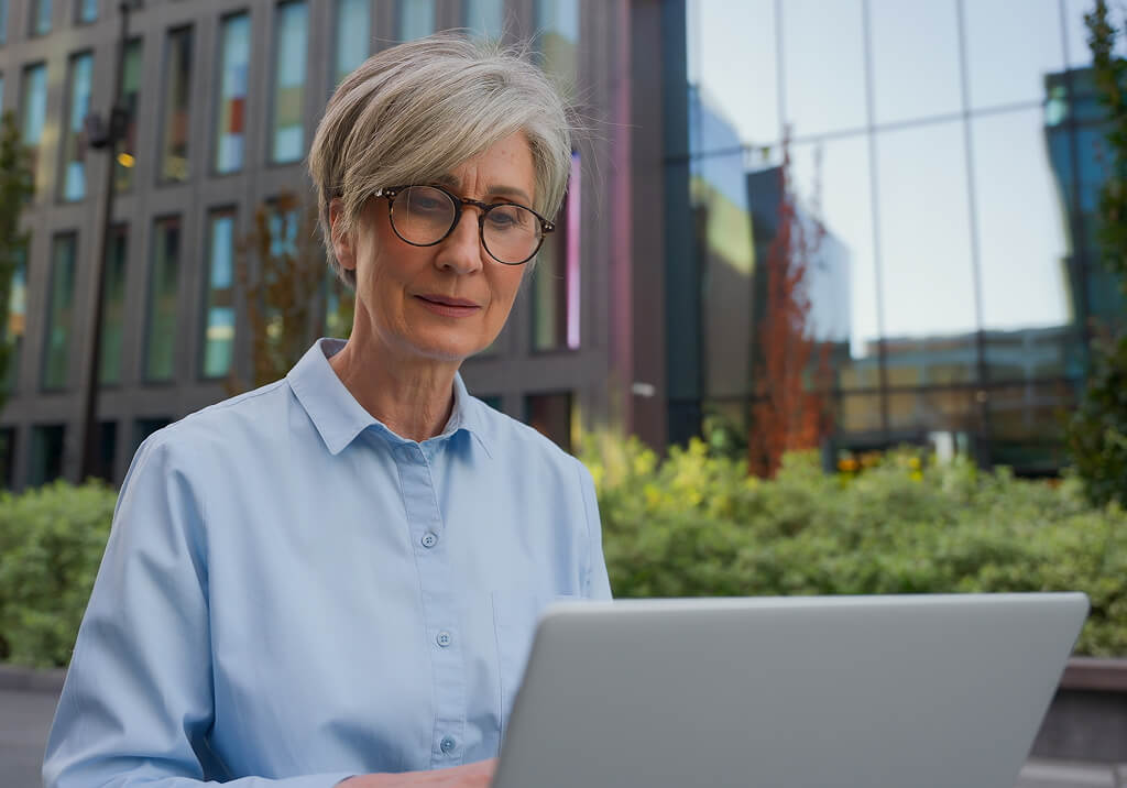 Older woman wearing glasses using a laptop outdoors in front of a modern office building.